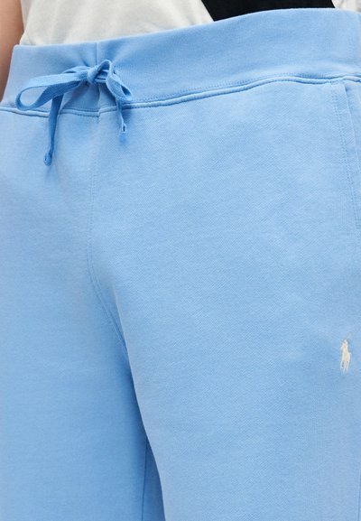 Ανοιχτό μπλε φόρμα με ραβδωτό κορδόνι στη μέση και μικρό λευκό κεντητό λογότυπο στο μπροστινό μηρό, φορεμένη με μαύρο και λευκό τοπ.