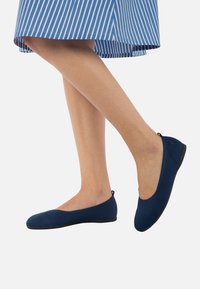 Ballerines bleu marine en tissu texturé, au design minimaliste, avec un bout arrondi, un profil bas et un petit détail de talon. Portées avec une jupe rayée bleue.