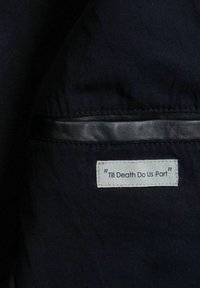 Melna auduma gluda faktūra ar baltu etiķeti. Etiķetē melnā krāsā rakstīts "Till Death Do Us Part".