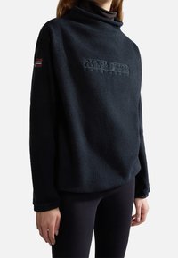 Pullover in pile nero con colletto alto, caratterizzato da un tessuto testurizzato e un logo in rilievo sul fronte, con un piccolo dettaglio a bandiera sulla manica.