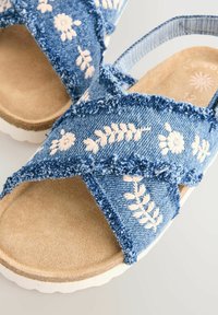 Sandali in denim con cinturini incrociati, tessuto blu con ricami floreali bianchi, soletta morbida color tan e suola in gomma bianca testurizzata.