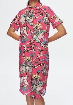 Femme portant une robe-chemise rose à manches courtes, longueur genou, avec de grands motifs floraux et feuillus multicolores. Bras pliés à la taille.