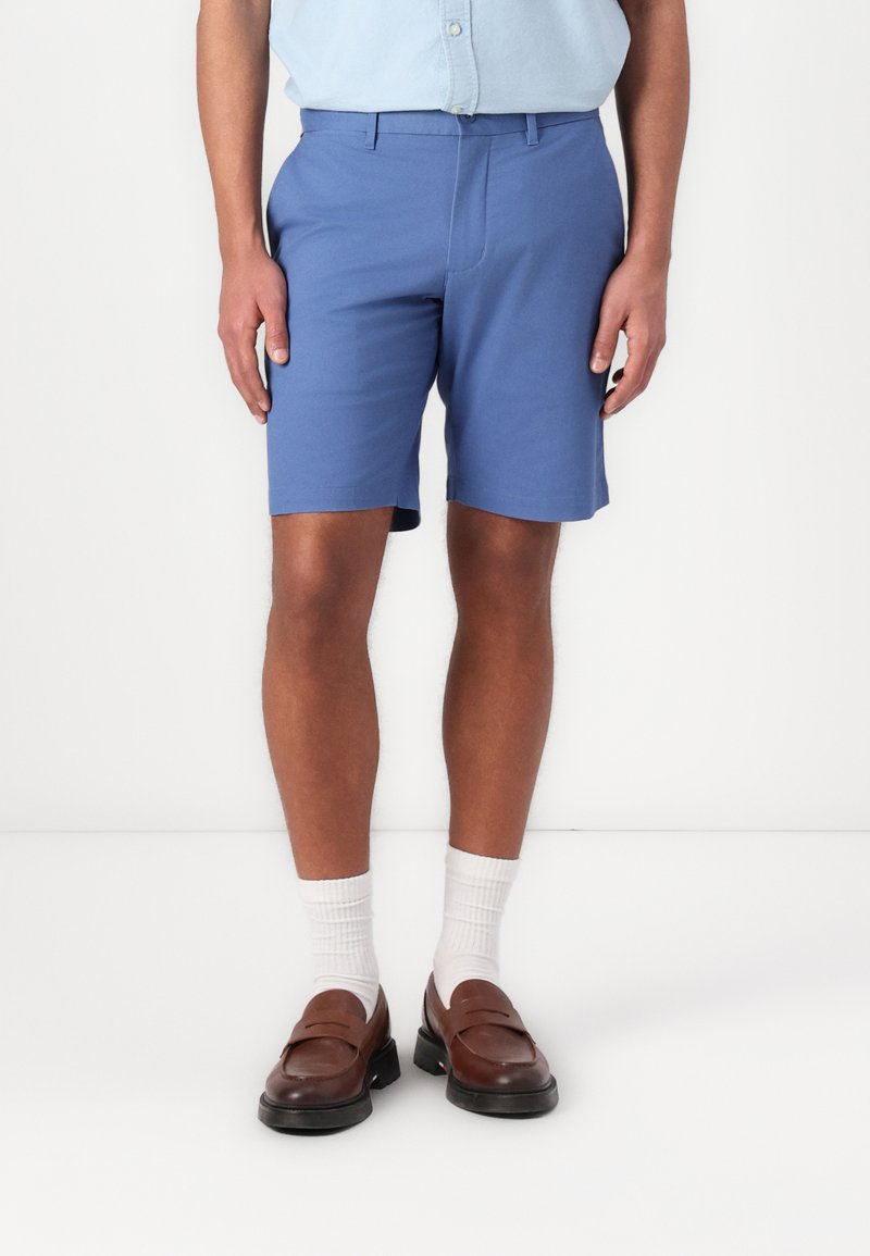 Shorts bleus en tissu lisse, avec une coupe droite, poches latérales, portés avec des mocassins marron et des chaussettes blanches de type crew.