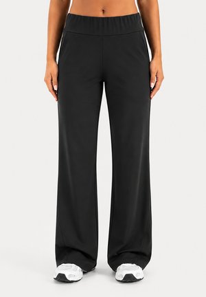 Pantalon noir ample avec taille élastique porté par une personne debout, accompagné de chaussures de sport blanches sur fond uni.