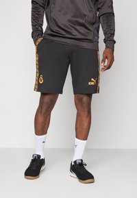 Czarne spodenki sportowe z pomarańczowymi akcentami i logo po bokach, połączone z białymi skarpetkami i czarnymi trampkami. Materiał wydaje się gładki i elastyczny.