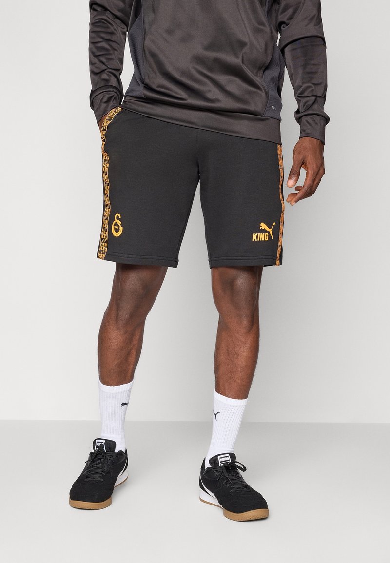 Czarne spodenki sportowe z pomarańczowymi akcentami i logo po bokach, połączone z białymi skarpetkami i czarnymi trampkami. Materiał wydaje się gładki i elastyczny.