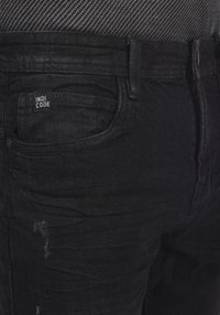 Jeans de mezclilla negra con una textura desgastada, que cuentan con una etiqueta de parche cosida en el bolsillo delantero, diseñados para un ajuste ajustado.