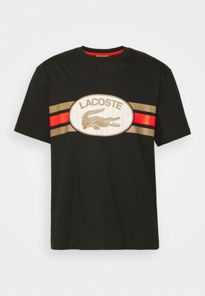 Czarna bawełniana koszulka z centralnym owalnym logo zawierającym napis "LACOSTE" oraz krokodyla, ozdobiona czerwonymi i złotymi poziomymi paskami.
