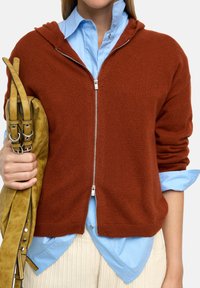 Rdzawoczerwony dzianinowy hoodie na zamek z kapturem, noszony na lekko niebieską koszulę. Model trzyma żółtą torbę z podwójnymi ramiączkami.