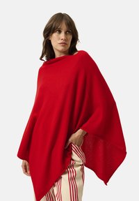 Poncho rosso con un ampio scollo drappeggiato e una vestibilità morbida. Realizzato in tessuto morbido e testurizzato. Abbinato a pantaloni a righe color crema.