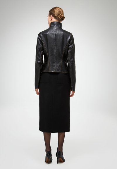 Nainen, jolla on punottu tukka, musta nahkatakki, pitkä musta hame, läpikuultavat sukkahousut ja mustat korkokengät, seisoo selin katsojaan.