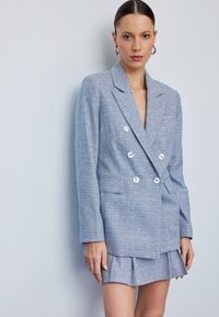 Blazer de doble botonadura en azul claro con tejido texturizado, botones blancos y un diseño ajustado. Combinado con una mini falda plisada a juego.