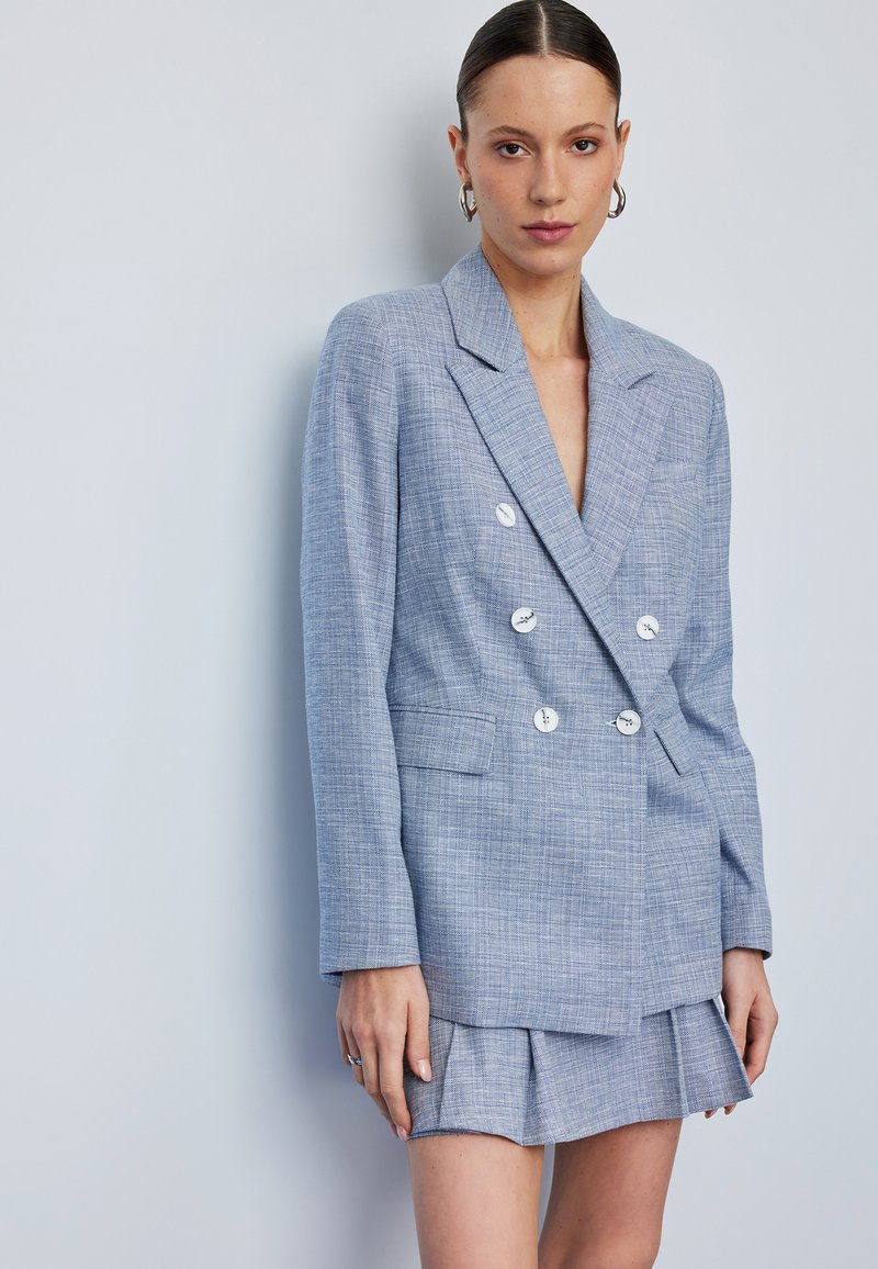 Blazer de doble botonadura en azul claro con tejido texturizado, botones blancos y un diseño ajustado. Combinado con una mini falda plisada a juego.