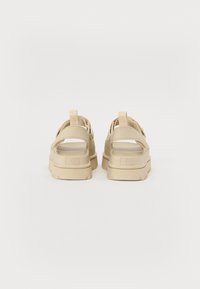 Beige sandaler med tykk såle, åpen hæl og stropper over foten. Teksturerte detaljer på overdelen med en løkke av stoff for enkel påkledning.
