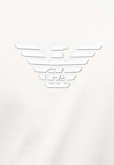 Λογότυπο αετού με στυλιζαρισμένα φτερά απλωμένα και ενσωματωμένα τα γράμματα "GA", τυπωμένο με λευκό χρώμα σε λευκό ύφασμα.