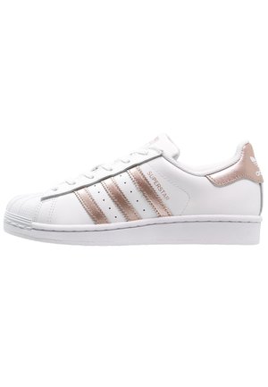 Hvid Adidas Superstar sneaker med rosenguld striber og hælkappe, gummiskal-tå og teksten "SUPERSTAR" på siden.
