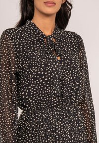 Robe noire avec un motif à pois crème. Manches longues semi-transparentes avec un col à nouer et une taille plissée pour plus de texture.