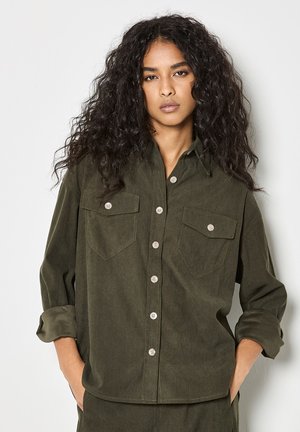 Olive green corduroy shirt with button closure, two chest pockets, rolled sleeves, and textured fabric. Classic design with subtle accents.