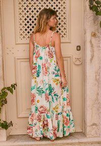 Robe maxi florale avec bretelles spaghetti, présentant une base blanche et des motifs floraux vibrants en rose, vert et jaune. Détail de ourlet volants.