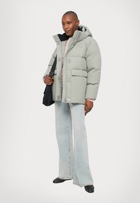 Lichtgrijze puffer jas met een capuchon, voorzien van grote voorzakken en een ritssluiting, gecombineerd met brede lichtblauwe jeans en zwarte schoenen.