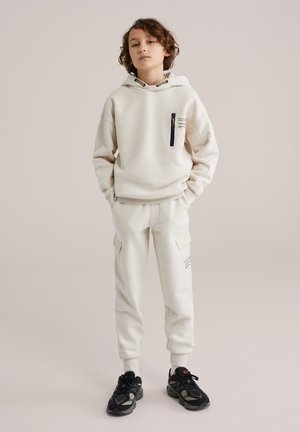 Ensemble sweat à capuche et pantalon couleur crème avec une poche zippée, des poignets côtelés et des poches latérales. Des baskets noires complètent le look.