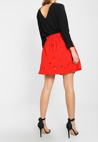 Top noir à manches longues avec un dos en V, associé à une jupe rouge ornée de motifs floraux, et des sandales à talons hauts noires.