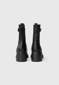 Um par de botas de couro pretas até ao tornozelo com saltos em bloco e correias com fivela, mostradas de costas sobre um fundo branco liso.