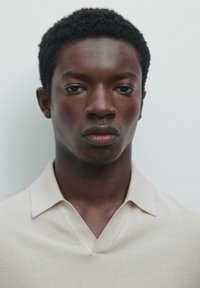 Giovane uomo nero con capelli ricci corti, indossa una camicia colletto beige chiaro, guarda dritto davanti a sé contro uno sfondo bianco semplice.