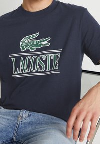 Man wearing navy blue Lacoste shirt with green crocodile logo and white outlined text, sitting with light blue jeans and hand resting on chair.