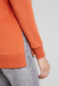 Sudadera naranja con puños y dobladillo acanalados, con aberturas laterales. Combinada con jeans gris claro, exhibiendo un look casual y cómodo.