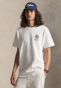 Junge Person mit langen lockigen Haaren, die ein weißes Ralph Lauren T-Shirt, weiße Hose, blaue "Ralph"-Kappe und eine Uhr trägt.