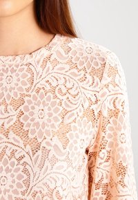 Pitsi toppi vaalean persikan värissä, jossa on kukkakuvio ja avoin kudontasuunnittelu, pyöreä pääntie ja pitkät hihat. Pehmeä rakenne, yksityiskohtainen kirjontakuvio.