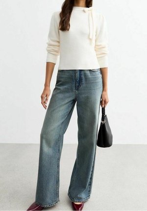 Mujer con un top de manga larga color crema con detalle de lazo, jeans de pierna ancha de talle alto, zapatos burdeos y sosteniendo un bolso negro.