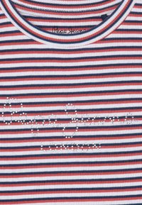 Koszulka w paski w kolorach czerwonym, białym i granatowym z prążkowaną fakturą. Posiada na froncie logo zapisane w stylu jubilerskim. Okrągły dekolt.