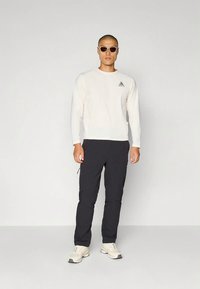 Mees, kes kannab valget pika varrukaga särki, musti pükse, beeže tossusid ja päikeseprille, seisab ühevärvilise helehalli tausta ees.