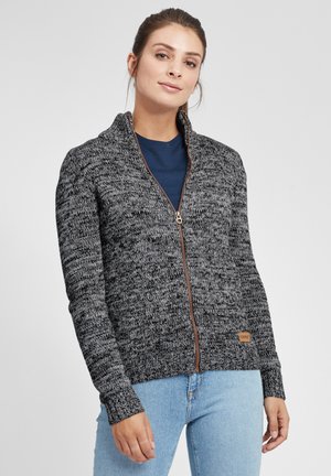 Sweater zippé gris en tricot avec des poignets côtelés, fermeture éclair marron et une petite étiquette de marque en bas. Porté sur une chemise bleu foncé.