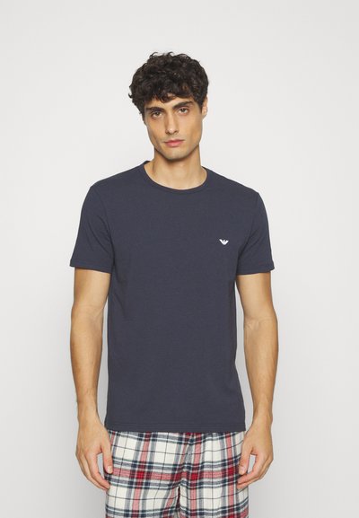 Navy blue cotton T-shirt with a round neckline and a small white logo on the left chest. Model is wearing plaid pajama pants.