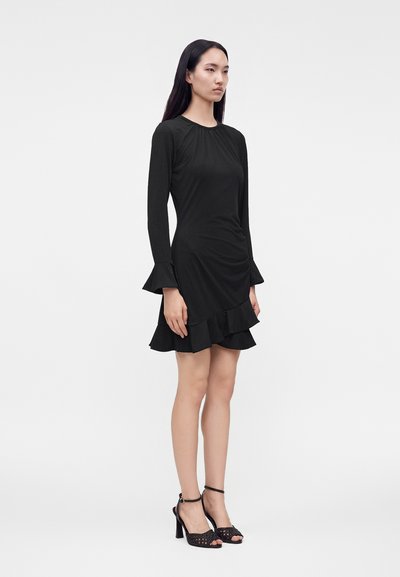 Femme portant une robe noire à manches longues avec des volants aux poignets et à l'ourlet, associée à des sandales noires à talons hauts, debout devant un fond uni.