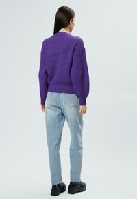 Lila gestrickter Pullover mit geripptem Muster, kombiniert mit hellblauen Jeans. Schwarze klobige Schuhe runden das Outfit ab.