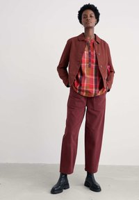 Raundpruun jakker koos kaelusega, nööbiga kinnitamine ja vastavad raundpruunid püksid. Kihtidega ülemine osa, millel on ruuduline mustriga punase, kollase ja rohelise tooni kombinatsioon. Mustad pahkluu saapad täiustavad riietust.