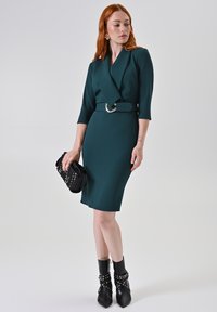 Teal getailleerde jurk met een wikkelontwerp, driekwart mouwen en een decoratieve riem. Gepaard met zwarte enkellaarsjes en een kleine tas.