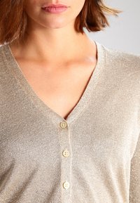 Kvinna som bär en beige glittrig V-ringad cardigan med tre synliga krämfärgade knappar, kort brunt hår och naturlig sminkning.