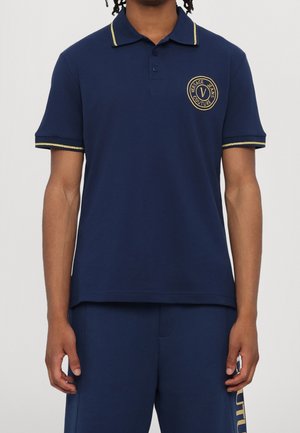 Polo bleu marine avec des bordures dorées sur le col et les manches. Présente un logo rond sur la poitrine. Confectionné en tissu mélangé de coton lisse.