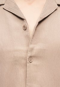 Μπεζ πλεκτό πουκάμισο με ρίγες, δύο ορατά κουμπιά και φαρδιά γιακά, που φοριέται ελαφρώς ανοιχτό στο λαιμό.