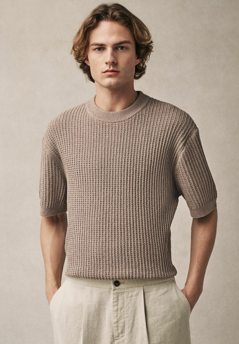 Suéter de punto beige de cuello redondo con mangas cortas y patrón texturizado; el modelo lleva pantalones de lino de color claro, con las manos en los bolsillos.