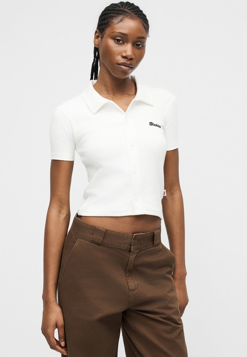 Giovane donna con capelli intrecciati che indossa una polo bianca corta e pantaloni marroni a vita alta, in piedi davanti a uno sfondo semplice.