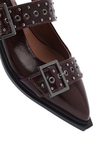 Zapato de charol burdeos con punta afilada, que presenta dos correas con hebilla adornadas con tachuelas plateadas y suela de goma negra.