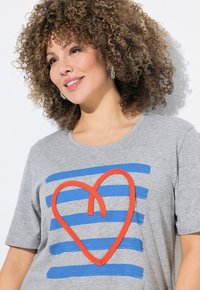 Camiseta de algodón gris con un diseño de corazón rojo de gran tamaño y rayas horizontales azules. Mangas cortas, escote redondo y textura suave.