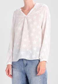 Blusa bianca a scollo a V in tessuto trasparente, con motivo a stelle su tutta la superficie. Maniche lunghe e vestibilità ampia, caratterizzata da una leggera texture.