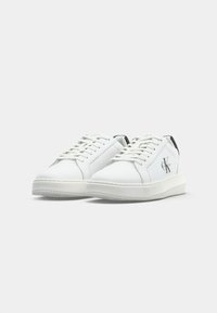 Witte leren low-top sneakers met veters, zwarte hieltabbladen en het Calvin Klein "CK" logo aan de buitenzijden, zij aan zij weergegeven.